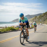 Two cyclists riding on a road with a scenic background of beach and mountains.